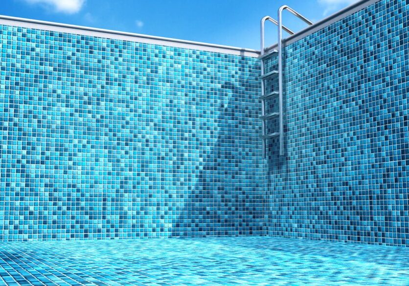 Empty swimming pool with blue mosaic tiles covering the walls and floor, metal ladder at the deep end, and a clear blue sky overhead.