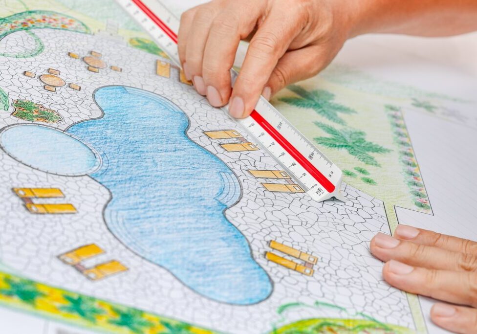 Hands using a scale ruler to measure a landscape plan featuring a pool, patio area, and surrounding foliage.
